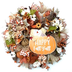 Puede incluir: Una corona con tem&aacute;tica oto&ntilde;al con un dise&ntilde;o de espantap&aacute;jaros y calabaza. La corona presenta un espantap&aacute;jaros con sombrero marr&oacute;n y una calabaza con el texto "Happy Fall y'all". Est&aacute; decorada con detalles en naranja, marr&oacute;n y blanco.