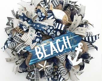 Puede incluir: Una corona decorativa con cintas de malla azul marino y beige con patrones náuticos que incluyen anclas, estrellas de mar y conchas. Un letrero de madera azul con el texto blanco BEACH está centrado, junto a un ancla blanca y un recorte de pez.