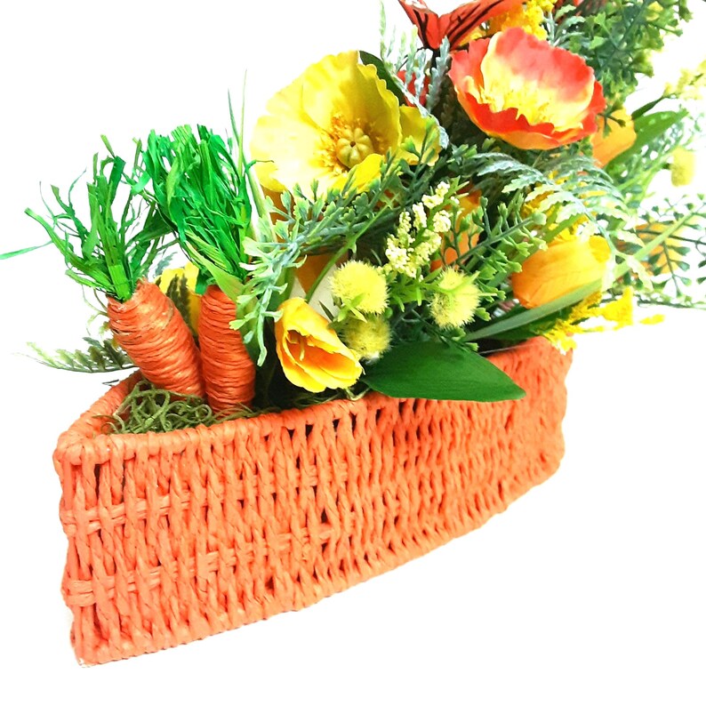 May include: An orange woven basket brimming with artificial flowers and foliage. The display features orange carrots, yellow tulips, and other bright blooms, creating a festive Easter or spring theme. The basket is made of woven material.