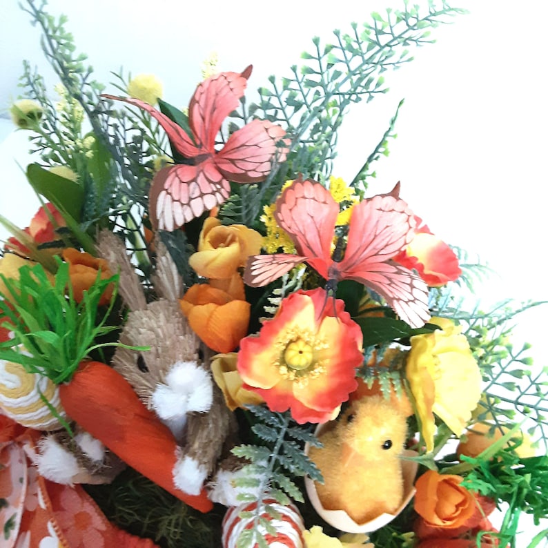 May include: An Easter wreath with a bunny, carrots, and a chick. The wreath features orange and yellow flowers, green foliage, and pink butterfly decorations. A festive spring decoration with a seasonal theme.