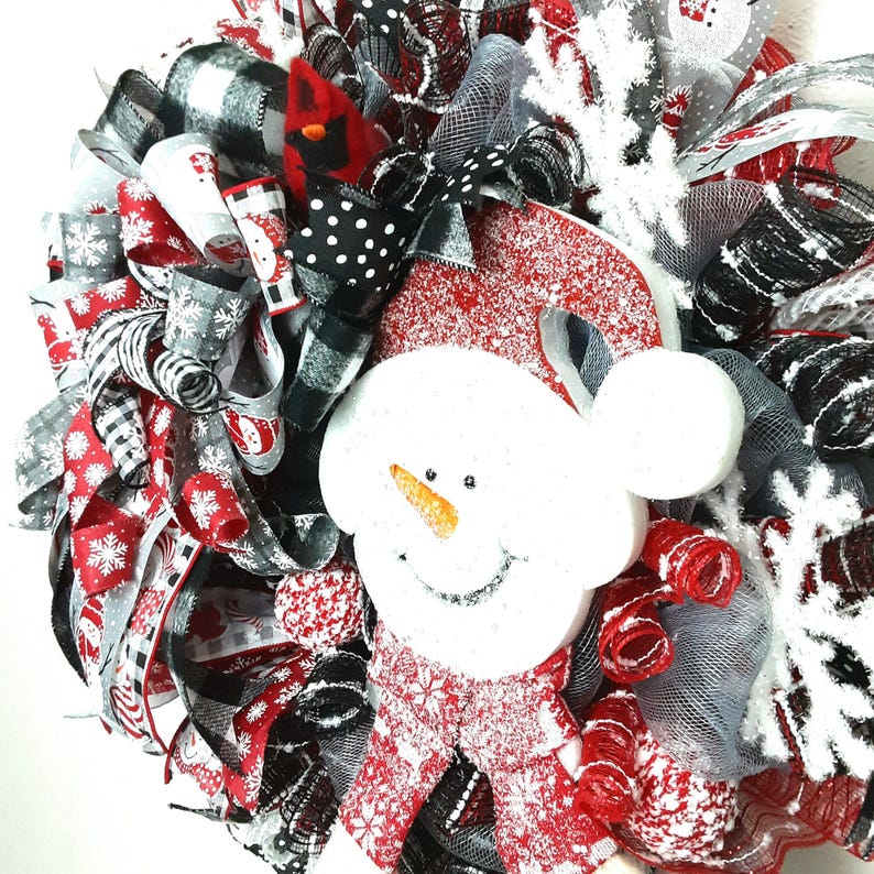 Puede incluir: Una corona navide&ntilde;a decorativa con un mu&ntilde;eco de nieve blanco central con nariz naranja, rodeado de cintas de malla rojas, negras y blancas, patrones de cuadros escoceses, estampados de copos de nieve y un peque&ntilde;o p&aacute;jaro cardenal rojo.