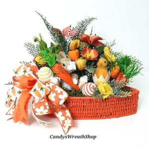 May include: An Easter basket arrangement featuring a bunny, chick, and decorative eggs. The basket is filled with colourful flowers, greenery, and a large orange bow with carrot and daisy patterns. The text "CandysWreathShop" is visible.