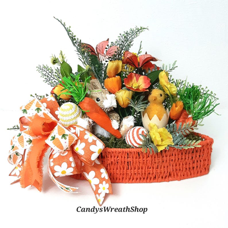May include: An Easter basket arrangement featuring a bunny, chick, and decorative eggs. The basket is filled with colourful flowers, greenery, and a large orange bow with carrot and daisy patterns. The text "CandysWreathShop" is visible.