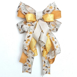 May include: A decorative bow featuring a bee-themed design. The bow has multiple layers of ribbon in beige, yellow, and gold. The beige ribbon is patterned with bees and floral elements. Yellow honeycomb and gold ribbons add contrast. The bow is suitable for crafts and decor.