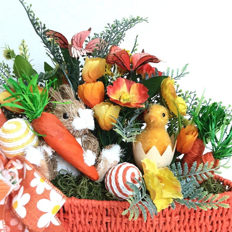 May include: An Easter basket filled with decorations. It includes a straw bunny, a fluffy yellow chick in an eggshell, faux carrots, and decorative eggs. The basket is filled with flowers and greenery, and has a bright orange woven design.