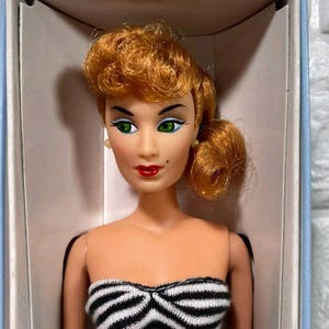 May include: A vintage doll with red hair styled in a bun, wearing a black and white striped swimsuit. The doll has green eyes and red lips. The doll is in a white box.