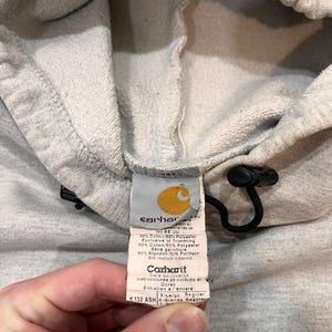Pu&ograve; includere: Un'etichetta di felpa con cappuccio Carhartt grigia con testo nero e un cordoncino nero. L'etichetta indica la composizione del tessuto come 50% cotone e 50% poliestere. L'etichetta include anche istruzioni di cura e informazioni sulle dimensioni: X-Large Regular, K132 ASH.
