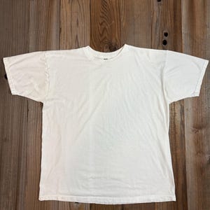 May include: A plain, off-white, short-sleeved t-shirt. The shirt is laid flat on a wooden surface, showcasing its simple design and neutral color. The t-shirt has a crew neck and a relaxed fit.