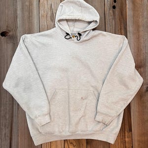 Pu&ograve; includere: Una felpa con cappuccio di colore grigio chiaro con una tasca a marsupio e un piccolo logo sulla tasca. La felpa ha un cordoncino.