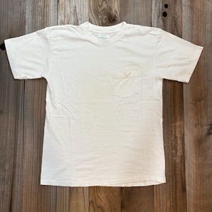 May include: A plain white t-shirt with a chest pocket. The short-sleeved shirt is made of a soft material and is laid flat on a wooden surface. The shirt has a crew neck.