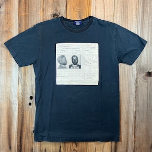 May include: Dark navy blue t-shirt featuring a black and white image of a document with two mugshot photos. The document has text and numbers. The t-shirt is displayed on a wooden background.