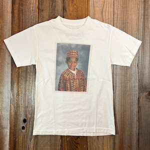 May include: White t-shirt with a printed image of a young boy wearing a patterned shirt and a hat. The boy is smiling and looking at the camera.