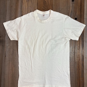 Peut inclure: Un t-shirt uni blanc cassé à manches courtes. Le t-shirt a un col rond et semble être fait d'un tissu doux et respirant. Le t-shirt est posé à plat sur une surface en bois.