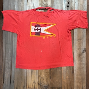 May include: A distressed red t-shirt featuring a graphic design. The design includes a white and yellow pennant shape with a central emblem and the letters "H I Y C" below. The shirt has a worn, vintage appearance.