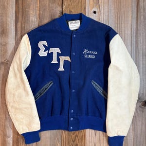 May include: A vintage blue and cream varsity jacket with the name "Harris Sluggo" embroidered on the front. The jacket features white lettering and cream-colored sleeves. The jacket has a blue collar, cuffs, and a button-up front.