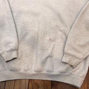 Pu&ograve; includere: Una felpa con cappuccio grigia con un piccolo logo arancione che dice "Carhartt" sul davanti. La felpa ha una tasca a marsupio e un cappuccio con coulisse.