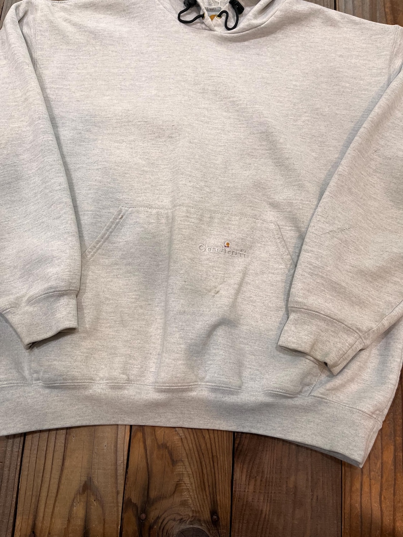 Pu&ograve; includere: Una felpa con cappuccio grigia con un piccolo logo arancione che dice "Carhartt" sul davanti. La felpa ha una tasca a marsupio e un cappuccio con coulisse.