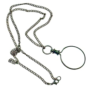May include: An antique-style magnifying glass pendant necklace. The necklace features a dark, chain with a circular magnifying glass attached. The magnifying glass is made of a dark metal and has a simple, elegant design. The chain has a clasp.