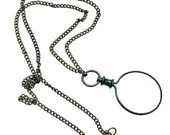 May include: An antique-style magnifying glass pendant necklace. The necklace features a dark, chain with a circular magnifying glass attached. The magnifying glass is made of a dark metal and has a simple, elegant design. The chain has a clasp.