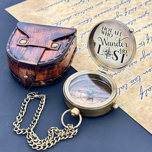 May include: A vintage brass compass with a chain and a leather case. The compass is open, displaying the needle and the text "NOT ALL WHO WANDER ARE LOST". The leather case is brown with a flap closure. A chain is attached to the compass.