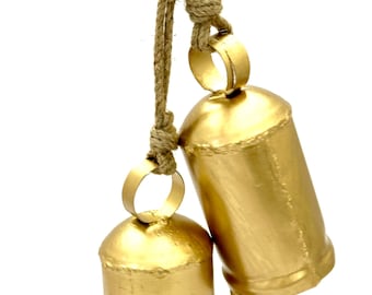 May include: Two gold-colored metal bells hanging from a natural rope. The bells have a cylindrical shape with a rounded top and a small loop for hanging. The rope is knotted at the top.