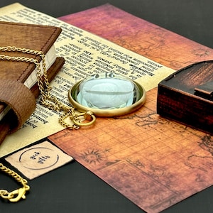 May include: A gold-colored pendant necklace with a clear, round glass orb. The pendant is set in a gold frame and hangs on a delicate gold chain. The necklace rests on a vintage map and a leather-bound book.