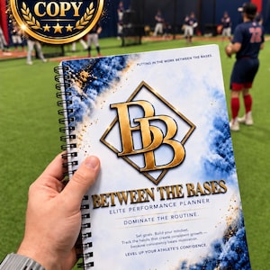 May include: A spiral-bound performance planner titled Between the Bases with a gold BB logo on a white cover featuring blue and gold smoke-like graphics. The text reads: Putting in the work between the bases, Elite Performance Planner, Dominate the routine.
