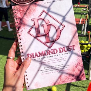 May include: A spiral-bound performance planner with a pink and white marble design. The cover features the text "DIAMOND DUST ELITE PERFORMANCE PLANNER", "EARN YOUR DIAMOND IN THE DUST", "DOMINATE THE ROUTINE", and "Set goals. Build your mindset."