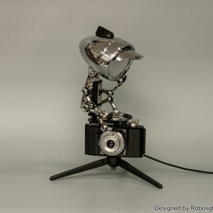 May include: A chrome robot lamp with a camera head, holding a vintage camera on a tripod. The lamp is designed by Robolight2018.