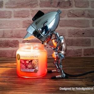 May include: A silver and copper robot lamp with a chrome head holding a glass candle jar. The candle jar is labeled "Yankee Candle Fresh Cut Roses".