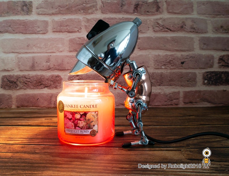 May include: A silver and copper robot lamp with a chrome head holding a glass candle jar. The candle jar is labeled "Yankee Candle Fresh Cut Roses".