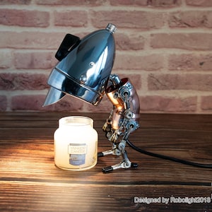 May include: A steampunk-style lamp made from repurposed metal parts, with a chrome-plated headlight for a shade. The lamp is illuminating a small glass jar candle with the label "Yankee Candle".