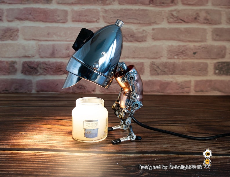 May include: A steampunk-style lamp made from repurposed metal parts, with a chrome-plated headlight for a shade. The lamp is illuminating a small glass jar candle with the label "Yankee Candle".