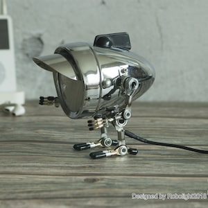 May include: A chrome-plated bicycle headlight lamp with a black visor, mounted on a metal stand with adjustable legs. The lamp is connected to a black wire.