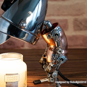 May include: A steampunk-style lamp with a chrome-plated headlight and copper pipe accents. The lamp is positioned as if it is looking down at a white candle jar with the label "Yankee Candle".