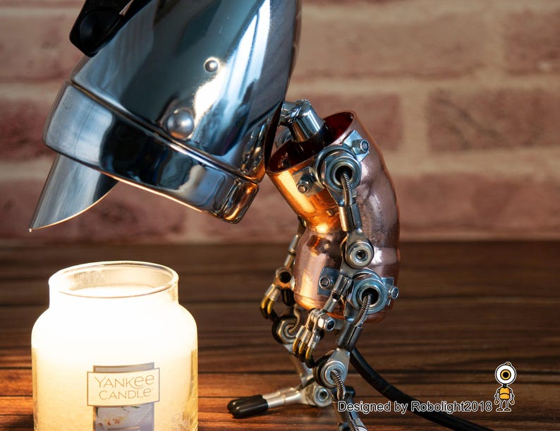 May include: A steampunk-style lamp with a chrome-plated headlight and copper pipe accents. The lamp is positioned as if it is looking down at a white candle jar with the label "Yankee Candle".