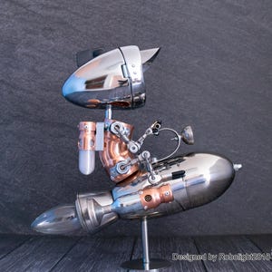 May include: A metallic robot sculpture featuring a chrome head and a rocket-shaped body made of polished silver and copper components. The robot is mounted on a thin metal stand and includes the text Designed by Robolight2018 with a small robot icon.