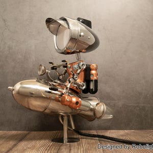 May include: A metallic robot-shaped lamp featuring a chrome finish, copper-colored accents, and a head shaped like a visor-covered light. The robot is mounted on a rocket-shaped base with a black power cord and a small logo reading Designed by Robolight2018.