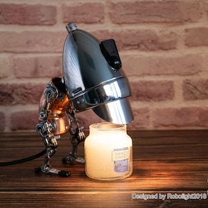 May include: A steampunk-style lamp with a chrome lampshade and copper and silver mechanical arms, illuminating a white candle jar with the text "Yankee Candle" on the label. The lamp is on a wooden surface.