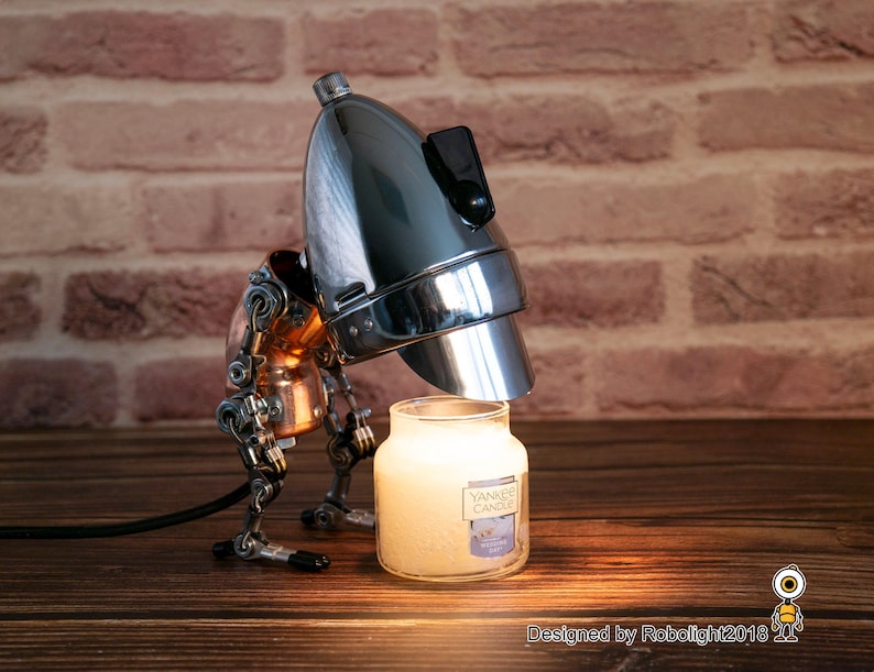 May include: A steampunk-style lamp with a chrome lampshade and copper and silver mechanical arms, illuminating a white candle jar with the text "Yankee Candle" on the label. The lamp is on a wooden surface.