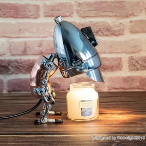 Könnte beinhalten: Eine Steampunk-Lampe aus Chrom und Kupfer mit einer einzigen Glühbirne. Die Lampe ist roboterförmig mit einem einzigen Bein und zwei Armen. Die Lampe leuchtet eine Glas-Glockenglocke mit dem Etikett "Yankee Candle" an.
