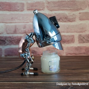 May include: A chrome and copper steampunk-style robot lamp with a black cord, holding a white candle jar with the text "Yankee Candle" on the label. The robot is standing on a wooden surface.