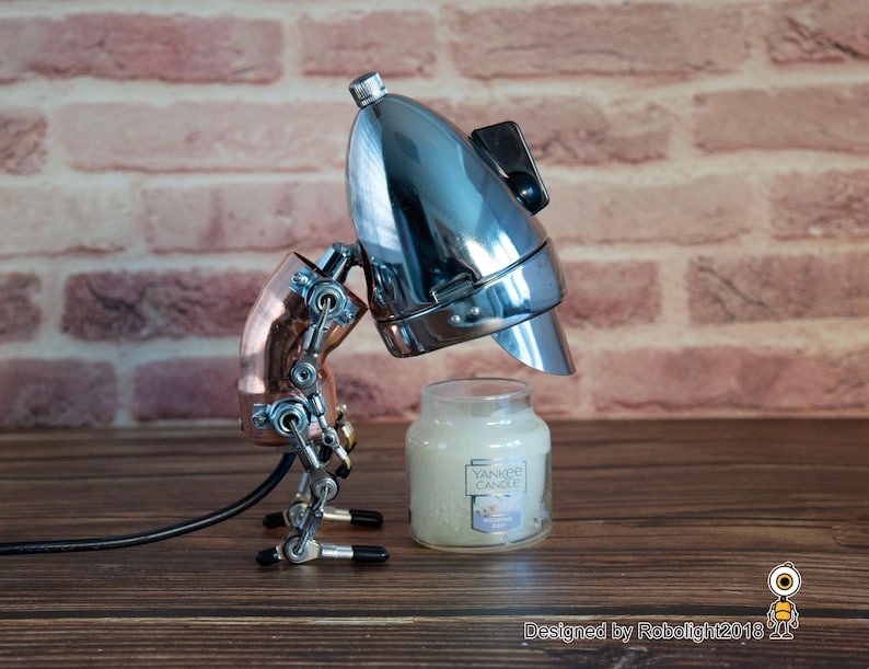 May include: A chrome and copper steampunk-style robot lamp with a black cord, holding a white candle jar with the text "Yankee Candle" on the label. The robot is standing on a wooden surface.