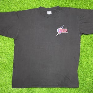 Peut inclure: Un t-shirt noir à manches courtes avec un petit motif sur la poitrine gauche. Le motif représente un emblème avec une épée et un bouclier accompagné du texte The Legend of Zelda Ocarina of Time en lettres rouges et blanches. Col rond.