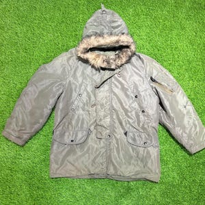 May include: A sage green winter parka featuring a hood with a faux fur trim, a front zipper closure with a storm flap, two large flap pockets at the waist, and a zippered utility pocket on the left sleeve. The jacket has a smooth, water-resistant finish.
