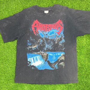 May include: A black short-sleeve t-shirt with a faded wash effect. The front features a graphic print with the red text Amorphis above a blue and black landscape illustration depicting a waterfall, rocky mountains, and a large axe in the foreground.