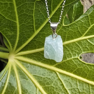 May include: A raw, pale blue aquamarine crystal pendant suspended from a delicate silver-toned metal chain. The stone features an irregular, unpolished, and translucent texture, held securely by a simple silver-colored metal bail at the top.