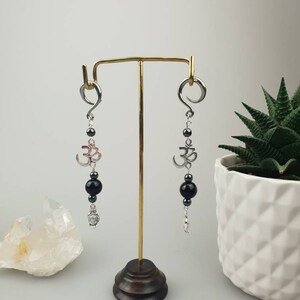 May include: A pair of dangle earrings with silver-tone hoops. Each earring features a silver Om symbol, black beads, and a small charm. The earrings are displayed on a gold-colored stand, with a white background.