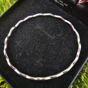 May include: A silver-colored, hammered-style bangle bracelet. The bracelet has a wavy, textured design and is displayed in a black box. The box is surrounded by green moss. The text on the card says "act with chemicals, creams and perfumes, great with the same love and care it was crafted with."
