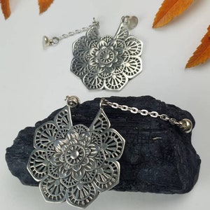 May include: Two silver-colored, flower-shaped tea infusers with intricate openwork designs. Each infuser is connected to a small chain and a round stopper. The infusers rest on a dark, textured surface, with orange leaves in the background.